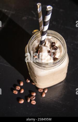 Un bocal en verre contient une boisson crémeuse au café surmontée de grains de café et de pailles rayées, placé sur une surface sombre avec des grains dispersés améliorant le contraste. Banque D'Images