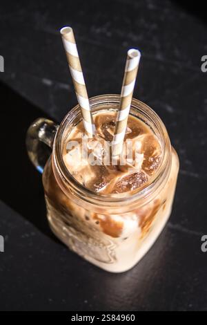 Un pot mason en verre rempli de café glacé et de glaçons repose sur une surface sombre. La lumière du soleil crée des ombres nettes, accentuant le contraste de la boisson. Banque D'Images
