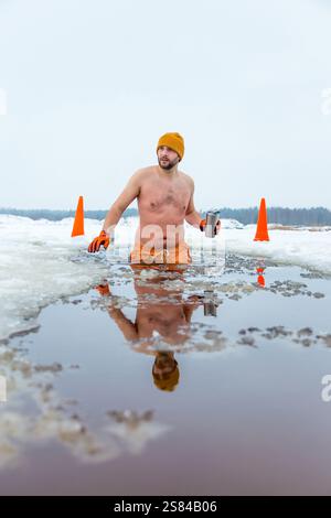 Un homme en short orange et bonnet tient la taille profondément dans l'eau glacée, tenant une tasse en métal. Neige, glace, cônes de circulation et toile de fond boisée complètent le Banque D'Images