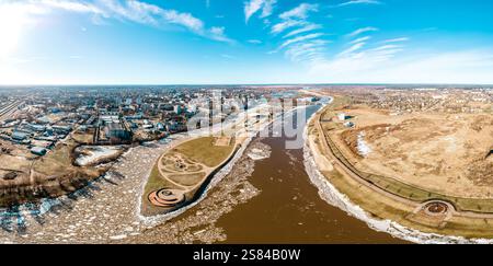 Vue aérienne panoramique d'une ville divisée par une rivière partiellement gelée, avec des bâtiments urbains, des champs ouverts et une structure en spirale circulaire près du Banque D'Images