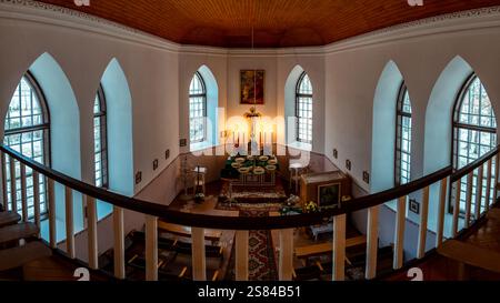 Vue depuis le balcon sur l'intérieur d'une église avec plafond en bois, fenêtres cintrées, bancs, moquette ornée, autel avec bougies et crucifix. Banque D'Images
