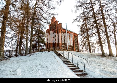 Une petite chapelle de style gothique avec des fenêtres cintrées et une entrée pointue se trouve au sommet d'une colline enneigée, entourée de grands arbres sans feuilles et de lumière du soleil. Banque D'Images