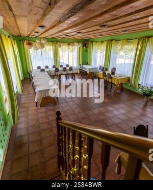 Salle à manger rustique avec poutres apparentes, murs à pois verts, grandes fenêtres aux rideaux jaunes et tables soigneusement disposées avec nappe Banque D'Images
