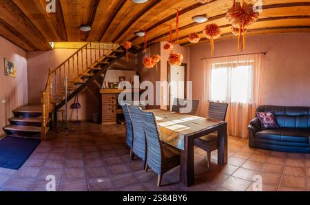 Chambre confortable avec plafond en bois, poutres apparentes, cheminée en briques, chaises en osier, canapé en cuir noir, et la lumière naturelle à travers des rideaux roses. Banque D'Images
