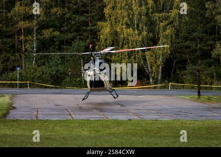 Un hélicoptère atterrit sur un héliport pavé bordé d'herbe et de forêt dense. Trois individus, dont deux en gilets de haute visibilité, sont présents à proximité Banque D'Images