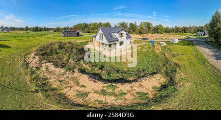 Une maison moderne de deux étages avec de grandes fenêtres et un toit sombre, entourée d'herbe et de végétation clairsemée, avec une dépression sablonneuse au premier plan. Banque D'Images