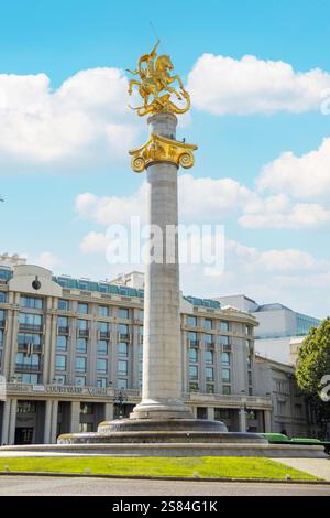 Le Freedom Monument à Tbilissi, Géorgie, présente une statue dorée de George au sommet d'une haute colonne, entourée de bâtiments modernes et d'un ciel clair. Banque D'Images