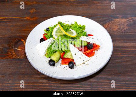 Salade colorée avec légumes verts frais, tomates mûres, concombre, olives et fromage feta présentés sur une assiette blanche. Une tranche de citron ajoute un refr Banque D'Images
