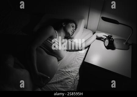 Une femme enceinte se couche dans son lit et tient un réveil. Banque D'Images