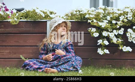 Fille contemplative en robe d'été, pieds nus parmi les fleurs blanches, tenant des framboises fraîches près d'une clôture en bois rustique Banque D'Images
