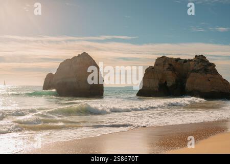 De majestueuses formations rocheuses s'élèvent de l'océan tandis que de douces vagues se jettent sur le rivage sablonneux sous un ciel doux et lumineux. Banque D'Images