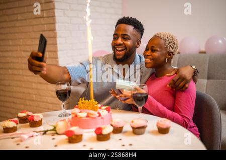 Heureux jeune couple célébrant l'anniversaire de la femme à la maison. Ils embrassent et prennent selfie. Banque D'Images