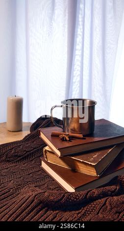 Tasse en métal sur pile de livres vintage avec des bâtons de cannelle sur une couverture tricotée marron doux, bougie brillante doucement en arrière-plan pour une ambiance hivernale. Banque D'Images