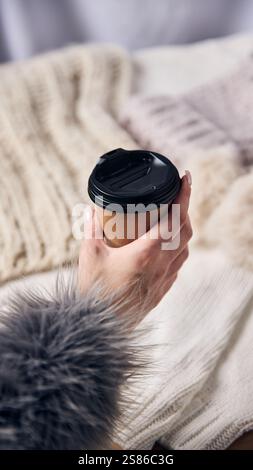 Main tenant une tasse de café avec manchette en fourrure douce dans un cadre confortable, reflétant les matins chauds d'hiver et les moments de vacances sereins. Banque D'Images