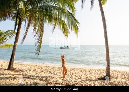 Jeune femme blonde debout sous les palmiers et profitant du soleil du matin à la plage tropicale de Maenam sur Koh Samui, Thaïlande. Banque D'Images