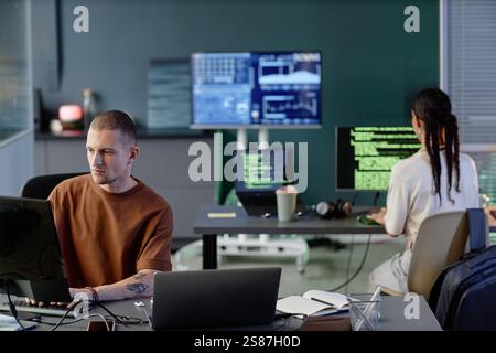 Plan grand angle de programmation masculine sur ordinateur au bureau travaillant avec des collègues dans une pièce spacieuse du bureau moderne de l'entreprise INFORMATIQUE, capturé sur de nombreux écrans avec des lignes de code Banque D'Images