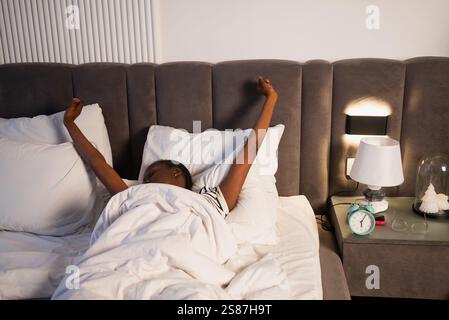 La femme s'étire dans son lit après le réveil, avec ses bras levés au-dessus de sa tête et une expression paisible sur son visage, profitant du début d'une nouvelle journée Banque D'Images