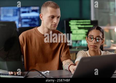 Plan moyen d'un programmeur INFORMATIQUE senior masculin examinant le code sur l'ordinateur portable de ses collègues tout en travaillant en collaboration au bureau Banque D'Images