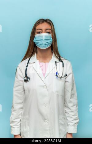 Vertical jeune femme médecin portant une blouse de laboratoire et un masque facial de protection debout sur fond bleu clair, représentant les soins de santé, la protection et Banque D'Images