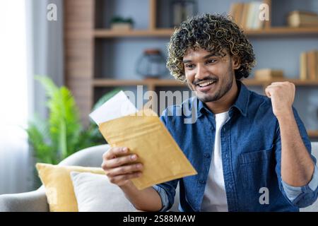 Homme réussi et joyeux assis sur le canapé dans le salon, tenant la lettre d'enveloppe postale dans les mains, célébrant la victoire et les résultats réussis obtenus dans notifié . Banque D'Images