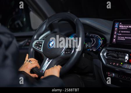 Mains femme saisissant le volant BMW iX3-M à l'intérieur de la voiture électrique avec tableau de bord éclairé et écran d'infodivertissement la nuit. Suède. Banque D'Images