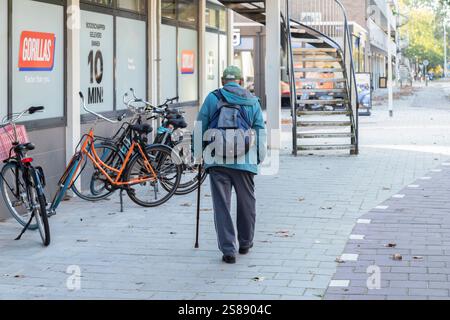 Vieil homme avec sac à dos et promenades en canne laborieuses sur le trottoir d'une rue commerçante déserte dans une banlieue d'Amsterdam. Banque D'Images