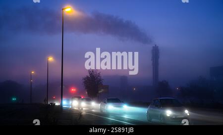 Conduite dans les embouteillages aux heures de pointe - trafic dans la ville illuminée dans des conditions humides et pluvieuses avec une mauvaise visibilité avec des cheminées industrielles. Banque D'Images