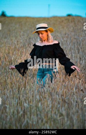 Une femme blonde se promène dans un champ de blé doré pendant la soirée. Elle est habillée d'un haut noir et d'un Jean bleu. Le coucher du soleil jette Banque D'Images