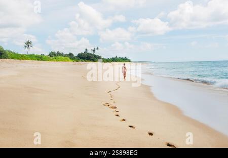 Jeune femme vêtue de vêtements d'été légers marchant pieds nus laissant des empreintes sur le sable sur l'océan Indien Tangalle solitaire cococotiers plage sur SR Banque D'Images