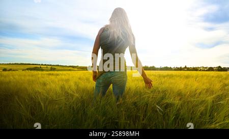 Femme agriculteur en jeans et t-shirt relaxe dans le champ de la nature au coucher du soleil - tient la main aux oreilles de blé ou d'orge - son heureux et équilibré. Banque D'Images
