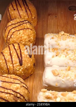 Diverses friandises sucrées sont habilement disposées sur un plateau en bois. Les boules rondes sont arrosées de chocolat, tandis que les barres sont enrobées de glaçage crémeux Banque D'Images
