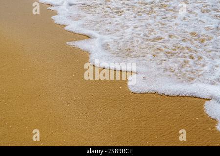 Gros plan d'une vague douce qui tourne doucement à la plage de sable sous un ciel bleu clair avec des nuages Banque D'Images