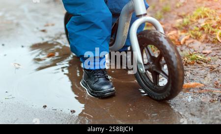 Un enfant roule sur un vélo d'équilibre à travers un chemin humide et boueux avec des flaques d'eau. L'image capture un moment d'enfance ludique et insouciant en profitant de l'outdoo Banque D'Images