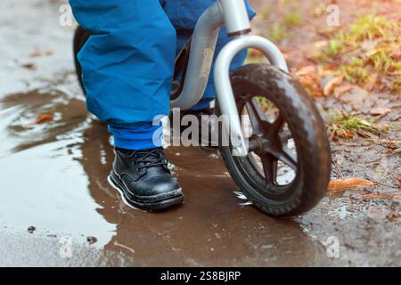Un enfant roule sur un vélo d'équilibre à travers un chemin humide et boueux avec des flaques d'eau. L'image capture un moment d'enfance ludique et insouciant en profitant de l'outdoo Banque D'Images