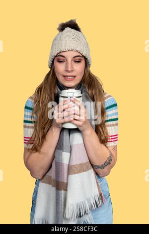 Femme souriante portant des vêtements d'hiver tenant la boisson chaude sur fond jaune Banque D'Images