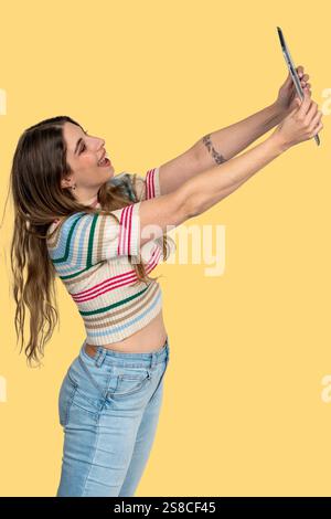 La jeune femme prend joyeusement un selfie avec une tablette, profitant d'un moment d'expression de soi sur un fond jaune vif Banque D'Images