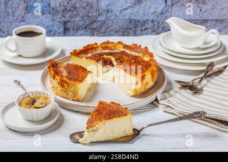 San Sebastian ou basque tranches de cheesecake brûlées sur l'assiette et sur la pelle à gâteau sur la table en bois blanc avec une tasse de café et de crème dans le fond de Banque D'Images