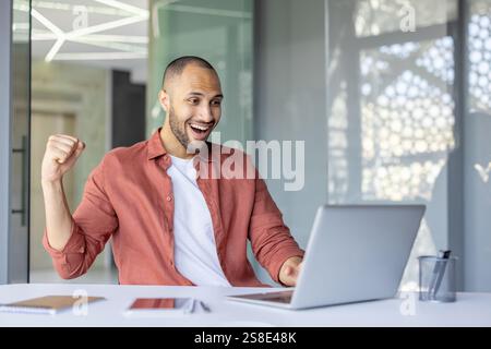Homme joyeux et réussi célébrant les résultats de réussite sur le lieu de travail, homme d'affaires dans des vêtements décontractés a reçu des nouvelles gagnantes et triomphantes en ligne, utilise un ordinateur portable. Banque D'Images