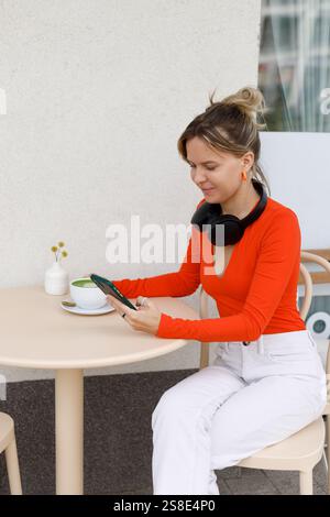 Une femme est assise dans un café en plein air à Varsovie, utilisant son smartphone et portant des écouteurs elle aime une tasse de matcha tout en s'engageant avec ses gadgets dans un Banque D'Images