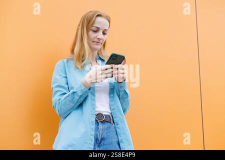 Une femme se tient contre un mur orange à Varsovie, utilisant intentionnellement son smartphone elle porte une chemise bleue et un Jean, se concentrant sur son écran, incarnant le mod Banque D'Images