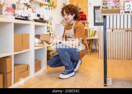 Un jeune employé de magasin, portant un tablier, squats pour organiser des boîtes en carton sur une étagère dans un petit magasin de magasin le cadre est chaud, avec des plantes et col Banque D'Images