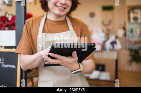 Le travailleur de détail souriant portant un tablier utilise une tablette à l'intérieur d'un magasin élégant L'arrière-plan présente des étagères floues et un décor, suggérant un ret moderne Banque D'Images