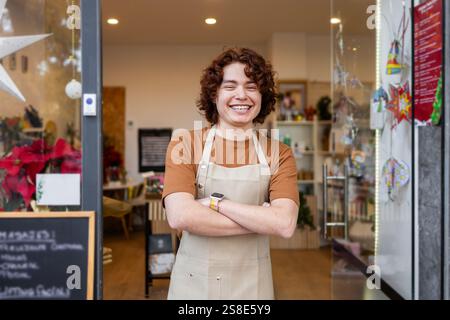Un jeune assistant de magasin joyeux dans un tablier se tient avec les bras croisés à l'entrée d'un magasin de produits biologiques, invitant les clients dans le confortable, e Banque D'Images