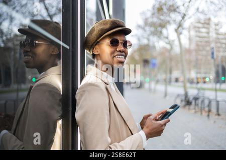 Une femme noire élégante dans un chapeau et des lunettes de soleil sourit tout en utilisant son smartphone elle s'appuie contre un mur de verre réfléchissant dans un environnement urbain extérieur Banque D'Images