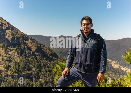 Un homme avec un sac à dos se tient confiant sur un sentier de montagne, entouré de collines luxuriantes et boisées sous un ciel clair et lumineux, Une image parfaite de l'extérieur Banque D'Images