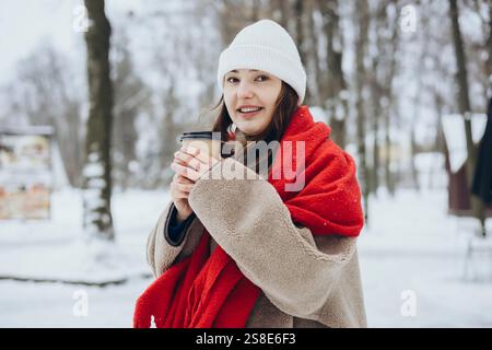 Une jeune femme en vêtements d'hiver tient une tasse à café enveloppée dans une écharpe rouge confortable, dans un paysage enneigé. Banque D'Images