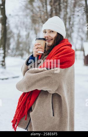 Une jeune femme joyeuse enveloppée dans un manteau confortable et une écharpe rouge, tenant une tasse à café dans un parc enneigé. Banque D'Images