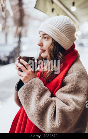 Une jeune femme réfléchie dans un bonnet blanc et une écharpe rouge tient une tasse chaude, regardant avec soin tout en étant entouré par une ambiance enneigée. Banque D'Images