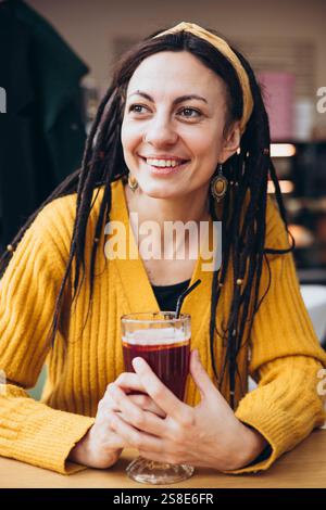 Une femme souriante avec des dreadlocks sirote un verre à travers une paille, assise dans un café confortable elle rayonne de joie et de chaleur, portant un pull jaune, créant un Banque D'Images