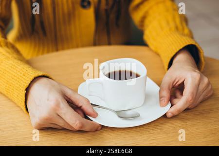Une scène chaleureuse d'une femme tenant une tasse de café sur une table en bois elle porte un pull jaune chaud, créant une atmosphère réconfortante et accueillante Perfe Banque D'Images